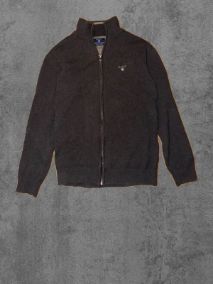 Mörkgrå ziptröja från Gant - Mörkgrå stickad ziptröja från Gant med hög krage och diskret logga på bröstet. Tröjan har hel dragkedja, ribbade muddar och är tillverkad i mjukt material. Perfekt för lager på lager och enkel att matcha. Passar S/M