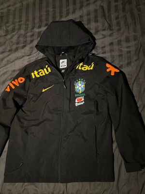 Nike Brasil svart vindjacka med huva - Ej använd. Riktigt bra kvalite. Mindre i stolek. Skulle säga att den är S/XS. Svart vindjacka från Nike med huva och dragkedja. Jackan har Brasilien CBF-logga, gula och orange sponsortryck samt Guaraná Antarctica-märke på bröstet. Perfekt för dig som gillar fotboll och vill ha en sportig stil. Riktigt fin