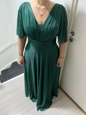 Grön långklänning med pärldetaljer - Elegant långklänning i mörkgrönt med v-ringning och vida, korta ärmar dekorerade med pärlor. Klänningen har ett markerat midjeband och ett mjukt, glittrande tyg som faller snyggt. Perfekt för speciella tillfällen.