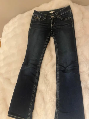 Mörkblå bootcut jeans med kontrastsömmar - Snygga mörkblå bootcut jeans med vita kontrastsömmar och klassiska fem fickor. Jeansen har låg midja och en liten detalj på ena fickan fram. Perfekta för en avslappnad men trendig look.