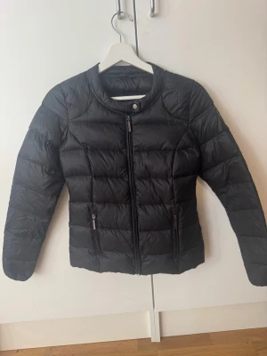 Svart pufferjacka med dragkedja - Snygg svart pufferjacka med quiltad design och rund krage. Jackan har dragkedja framtill, två fickor med dragkedja och knappdetalj vid halsen. Perfekt för övergångsväder och enkel att matcha med olika outfits. 