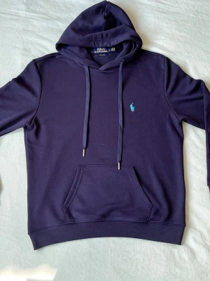 Marinblå hoodie från Polo Ralph Lauren - Snygg marinblå hoodie från Polo Ralph Lauren med klassisk huva och dragsko. Har en känguruficka framtill och den ikoniska broderade loggan i ljusblått på bröstet. Tillverkad i mjuk bomull för maximal komfort. Perfekt för en avslappnad och stilren look.