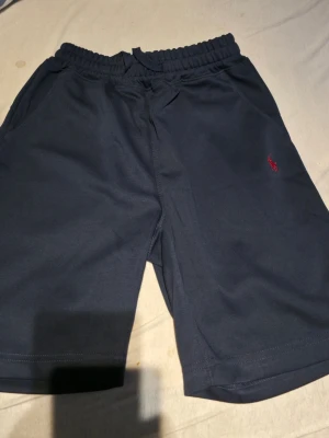 Mörkblå shorts från Ralph Lauren - Mörkblå shorts från Ralph Lauren med elastisk midja och snörning. Klassisk design med två sidofickor och den ikoniska röda loggan broderad på ena benet. Perfekta för sommaren och avslappnade dagar.