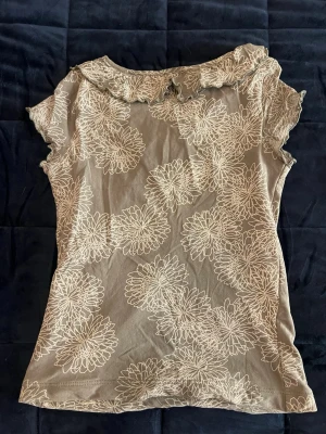 Beige blommig topp från Lindex - Supersöt beige topp från Lindex med vitt blommigt mönster och volangdetaljer vid den v-ringade halsen. Kortärmad och i mjukt material som känns skönt mot huden. Perfekt för dig som gillar en feminin och chill stil.