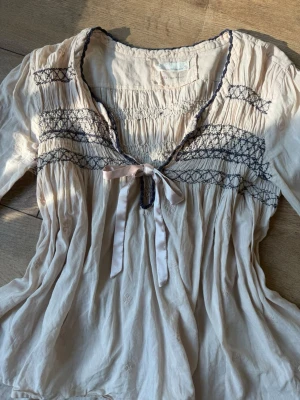 Beige blus med rosett från Odd Molly - Supersöt beige blus från Odd Molly med smockdetaljer och mörkblå broderier över bröstet. Blusen har en dekorativ rosett vid halsringningen och vågiga kanter. Tillverkad i ett tunt, mjukt material som ger en avslappnad och bohemisk känsla.