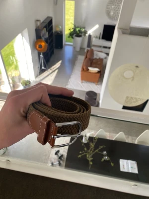 Brunt flätat bälte med läderdetaljer - Snyggt brunt flätat bälte med silverfärgat spänne och detaljer i brunt läder. Perfekt accessoar för att lyfta din outfit och ge en stilren touch. Bältet har en klassisk design och passar till både jeans och chinos.