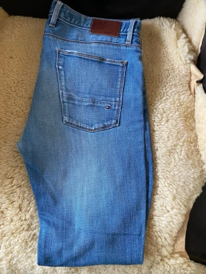 Blå raka jeans från Tommy Hilfiger - Säljer ett par klassiska blå jeans från Tommy Hilfiger med raka ben och normal midja. Jeansen har en snygg tvättad look, fem fickor och en liten logga på bakfickan. Perfekta till en avslappnad stil och passar till det mesta. Obs byxorna  är liten i storleken, se mått på bilderna
