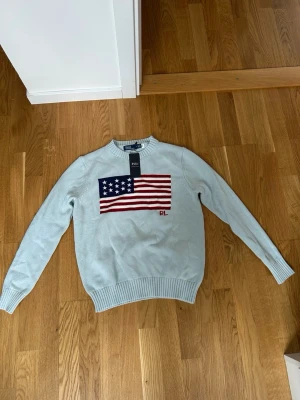 Ljusblå stickad tröja Polo Ralph Lauren - Ljusblå stickad tröja från Polo Ralph Lauren med amerikansk flagga broderad på bröstet och RL-detalj i rött. Tröjan har rund halsringning och ribbstickade muddar vid ärmslut och nederkant. Perfekt för dig som gillar klassisk preppy stil.