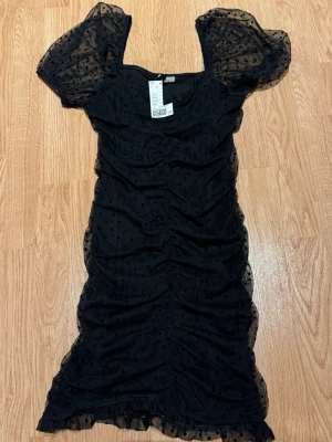 Svart prickig klänning från H&M - Svart kortklänning från H&M med puffärmar och transparent mesh med prickigt mönster. Klänningen har rynkade detaljer längs sidorna och volangkant nertill. Liten i storlek! 