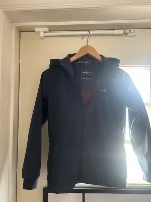 Mörkblå vindjacka från Sail Racing  - Snygg mörkblå vindjacka från Sail racing med huva och hel dragkedja framtill. Jackan har två sidofickor med dragkedja och diskret logga på bröstet samt på ärmen. Tillverkad i ett lätt och vindtätt material, perfekt för blåsiga dagar.