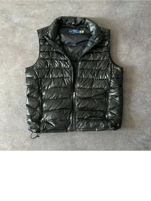 Svart dunväst från Polo Ralph Lauren - Snygg svart dunväst från Polo Ralph Lauren med hög krage och två dragkedjefickor. Västen har quiltad design och diskret logga på bröstet. Perfekt att slänga över en hoodie eller t-shirt för en chill och trendig look.