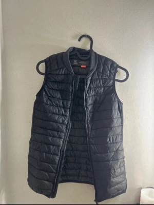 Svart dunväst från Haglöfs - Snygg svart dunväst från Haglöfs med quiltad design och diskret logga på bröstet. Västen har en hög krage i mjukt material och dragkedja framtill. Perfekt för lager-på-lager och håller dig varm under kyliga dagar.