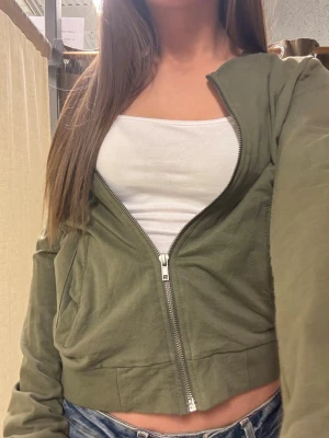 Olivgrön cropped hoodie med dragkedja - Snygg olivgrön cropped hoodie med dragkedja framtill och fickor på sidorna. Hoodien har en avslappnad passform och är tillverkad i mjukt bomullsmaterial. Perfekt att slänga på sig över ett linne eller t-shirt för en chill look.