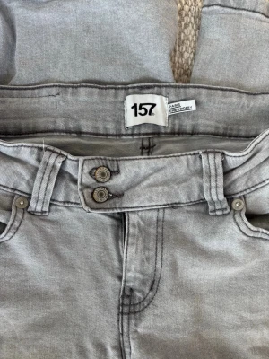 Grå jeans  - Säljer ett par grå jeans från lager 157 med snygg broderad detalj på bakfickan och dubbla knappar i midjan. Jeansen har klassisk femficksmodell och bälteshällor. Perfekta för dig som gillar en unik touch på dina jeans! Endast använda ett fåtal gånger.