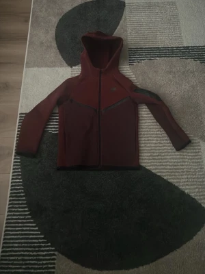 Vinröd hoodie från Nike - Snygg vinröd hoodie från Nike med hel dragkedja framtill och svarta detaljer längs bröstet och axlarna. Hoodien har en klassisk huva och lång ärm, perfekt för en sportig och avslappnad stil. Nike-loggan syns diskret på bröstet.