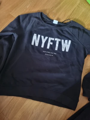 Svart sweatshirt NYFTW Gina Tricot - Cool svart sweatshirt från Gina Tricot med trycket 'NYFTW' i vitt framtill. Tröjan har rund halsringning och ribbade muddar vid ärmslut och nederkant. Perfekt för en avslappnad och trendig look.