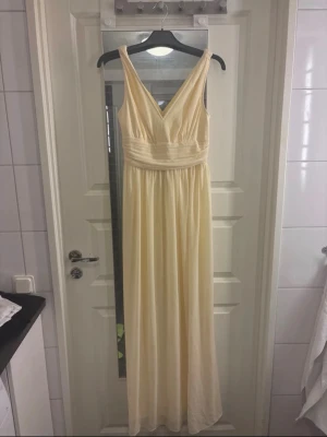 Ljus långklänning med v-ringning - Elegant långklänning i ljus beige med djup v-ringning både fram och bak. Klänningen har breda axelband och ett markerat midjeband som ger en snygg siluett. Tyget faller mjukt och ger en romantisk känsla, perfekt för sommarens speciella tillfällen.