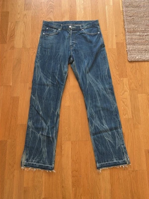 Blå raka jeans med rå kant - Säljer ett par blå denimjeans med rak passform och rå, fransig kant vid bensluten. Jeansen har klassisk femficksdesign, silverfärgade nitar och kontrastsömmar. Materialet är slitstarkt bomullsdenim med tydliga slitningar och tvättade detaljer.