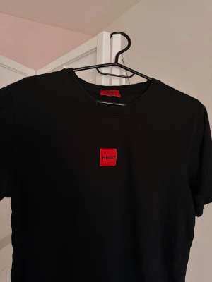 Svart t-shirt från Hugo med röd logga - Svart t-shirt från Hugo med en snygg röd logga på bröstet. Materialet är mjukt och känns skönt mot huden, t-shirten är tillverkad i bomull.