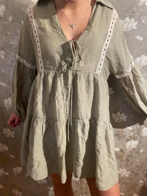 Ljusgrön kortklänning med spetsdetaljer - Supersöt ljusgrön kortklänning med volanger och vita spetsdetaljer längs ärmar och framtill. Klänningen har v-ringning med snörning och långa, lösa ärmar. Perfekt för en trendig och avslappnad look.