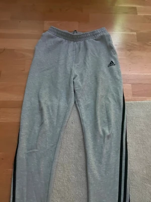 Gråa Adidas mjukisbyxor - Säljer ett par gråa mjukisbyxor från Adidas med klassiska svarta ränder längs sidorna och Adidas-logga på låret. Byxorna har resår i midjan och är gjorda i mjukt material, perfekta för chill dagar eller träning.