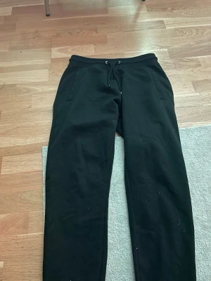 Svarta mjukisbyxor  - Ett par svarta mjukisbyxor med dragsko i midjan och fickor på sidorna. Byxorna har en bakficka och är tillverkade i ett mjukt och bekvämt material, perfekta för chill dagar. Passformen är avslappnad och benen är raka.