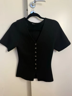 Svart v-ringad topp från Gina Tricot - Snygg svart topp från Gina Tricot med v-ringning och korta ärmar. Toppen har en figurnära passform och knappar framtill som detalj. Perfekt att styla med jeans eller kjol för en trendig look.