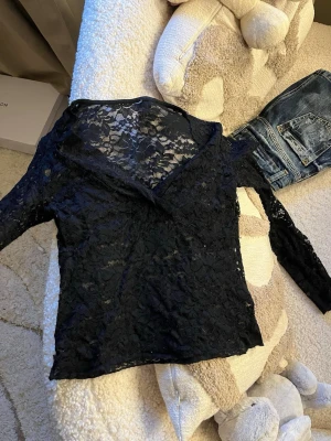 Svart spetsblus med v-ringning - Snygg svart blus i genombruten spets med v-ringad hals och långa ärmar. Blusen har ett blommigt spetsmönster som ger en elegant och trendig look. Perfekt att styla med jeans eller kjol för en cool outfit.