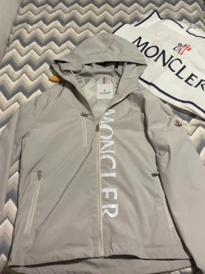 Beige vindjacka från Moncler - Snygg ljusgrå vindjacka från Moncler med huva och dragkedja framtill. Jackan har vita detaljer, stora Moncler-trycket längs dragkedjan och klassisk Moncler-logga på ärmen. Två sidofickor med dragkedja och en bröstficka. Denna moncler jackan har en liten defekt vid insidan av fickan som inte ens syns, annars är den jätte fin skicka. Sitter som M