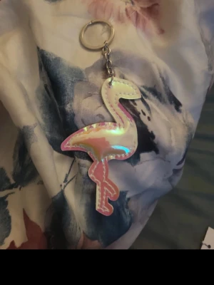 Holografisk flamingo nyckelring - Säljer en snygg holografisk nyckelring i form av en flamingo. Nyckelringen har skimrande yta i rosa, vitt och regnbågsskimrande toner. Perfekt att hänga på väskan eller nyckelknippan för en färgglad touch.