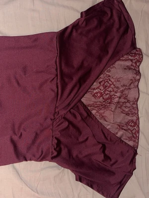 Vinröd topp med spets och twist - Snygg vinröd topp med korta ärmar och twistad front. Baksidan har transparent spetsdetalj i matchande färg som ger en unik look. Toppen är figurnära och passar perfekt till jeans eller kjol. Materialet känns mjukt och stretchigt.