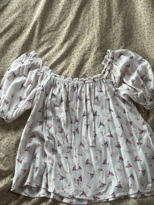 Vit blommig offshoulder blus - Söt vit blus med små blommor i rosa och lila toner och även orange. Modellen har puffiga offshoulder-ärmar med resår och en luftig passform. Perfekt för vår och sommar när du vill ha en romantisk look.