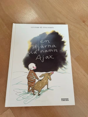 En stjärna vid namn Ajax - En illustrerad bok med vit hård pärm, skriven av Ulf Stark och illustrerad av Stina Wirsén. Omslaget visar ett barn i randig pyjamas och en hund mot en snöig bakgrund. Perfekt för dig som gillar känslosamma berättelser.
