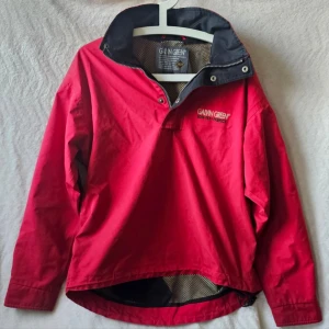 Röd vindjacka från Galvin Green - Snygg röd vindjacka från Galvin Green med svart krage och detaljer. Tillverkad i slitstarkt GORE-TEX-material som står emot vind och regn. Jackan har dragkedja och tryckknappar vid halsen samt meshfoder på insidan för extra komfort.