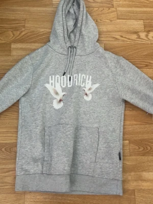 Grå HOODRICH hoodie med duvor - Snygg grå hoodie från HOODRICH med tryckta vita duvor och text på både fram- och baksidan. Stor magficka framtill och justerbar huva med dragsko. Materialet är mjukt och bekvämt, perfekt för en avslappnad streetwear-look.