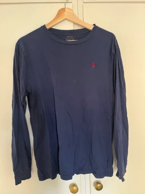 Blå långärmad topp från Polo Ralph Lauren - Mörkblå långärmad topp från Polo Ralph Lauren med rund hals och den klassiska röda broderade loggan på bröstet. Tillverkad i mjuk bomull som känns skön mot huden. Enkel och stilren design som funkar till det mesta.