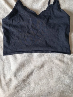 Svart croppad linnetopp med smala band - Snygg svart croppad linnetopp med smala axelband och en lätt vågig kant vid urringningen. Bild på skickas vid intresse.