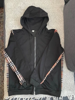 Svart hoodie med rutiga detaljer - Snygg svart hoodie med dragkedja och huva. Ärmarna har klassiska beige, röda och svarta rutiga paneler längs sidorna som ger en unik look. Mjuk bomullskänsla och ribbade muddar vid ärmslut och nederkant. Perfekt för dig som vill ha streetwear-vibe.