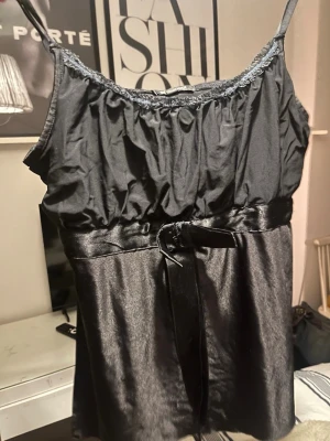 Svart satin linnetopp - Stilren svart linnetopp i satin med tunna axelband och spetsdetalj vid urringningen. Toppen har ett band i midjan som kan knytas för en snygg siluett. Perfekt för dig som gillar en elegant och enkel look.