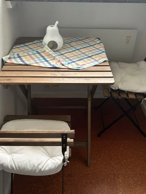 Balkongset med träbord och stolar - Säljer ett balkongset från IKEA med ett fyrkantigt träbord och två matchande stolar. Setet har vita, mjuka KUDDARNA dynor och svart metallstomme. Perfekt för mindre utrymmen och ger en mysig känsla till balkongen. Hämtas i Falköping 