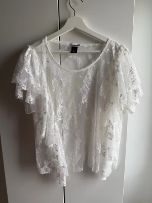 Ny Lindex vit topp i spets med volang. M - Ny Lindex vit topp i spets med volang. M  •Vit spetsblus med korta ärmar.  •Blusen har en rund halsringning och volanger på ärmarna och längs ena sidan framtill, vilket ger en feminin och lekfull touch.  •Spetsmönstret är detaljerat och blommigt, vilket skapar en elegant och luftig känsla.  •Blusen är genomskinlig och kan bäras över en topp eller linne för en komplett look. •Aldrig använd, endast tvättad.  🚭Djurfritt och rökfritt hem