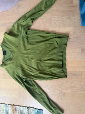 Grön stickad tröja Peak Performance. - Snygg grön stickad tröja från Peak Performance med V-ringning och långa ärmar. Tröjan har diskret logga på bröstet och ribbade muddar vid ärmslut och nederkant. Perfekt för lager på lager och enkel att matcha till olika outfits.