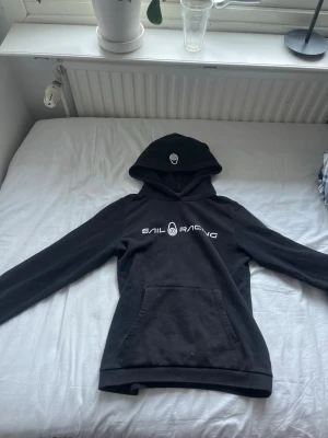 Svart hoodie från Sail Racing - Svart hoodie från Sail Racing med vit logga och text på bröstet. Klassisk känguruficka framtill och huva. Tillverkad i mjukt bomullsmaterial som är skönt att ha på sig. Perfekt för en avslappnad och sportig stil.