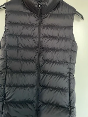 Svart dunväst Uniqlo med huva - Snygg svart dunväst med huva och dragkedja framtill. Västen har quiltad design med horisontella sömmar och två fickor med dragkedja. Perfekt för lager på lager och ger en sportig look. Materialet är glansigt och känns lätt men ändå varmt.