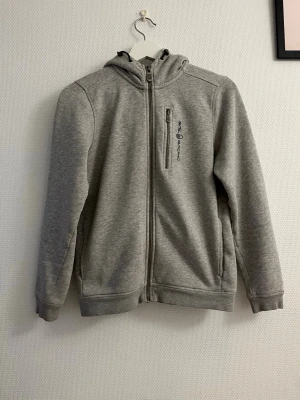 Grå hoodie från Sail racing - Snygg grå hoodie med dragkedja från Sail racing. Hoodien har huva, två sidofickor och en bröstficka med dragkedja. Tillverkad i mjukt material som känns skönt mot huden. Perfekt för dig som gillar en enkel och clean stil.