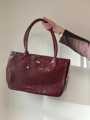 Stor röd (16) - Snygg vinröd handväska i croco-mönstrat konstläder med två rejäla handtag. Väskan har en stilren, fyrkantig form och en liten guldfärgad detalj framtill. Perfekt för dig som vill ha en trendig accessoar med lyxig känsla.