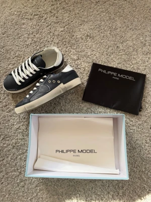 Philippe Model blåa lädersneakers - Snygga sneakers från Philippe Model i mörkblått läder med vita detaljer och vita skosnören. Skorna har en klassisk låg siluett, vit sula och metalliska öljetter. Perfekta för dig som gillar stilrena och exklusiva sneakers.