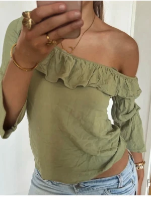Grön offshoulder topp med volang - Snygg ljusgrön offshoulder topp med volangdetalj längs halsringningen. Toppen har trekvartsärm och är croppad i modellen. Perfekt för dig som gillar en trendig och avslappnad stil.