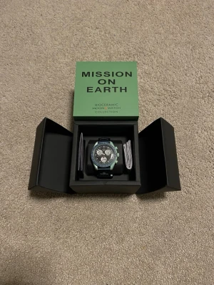Omega x Swatch MoonSwatch Mission On Earth - Säljer en Omega x Swatch MoonSwatch från Bioceramic MoonSwatch Collection. Klockan har en grön boett, svart urtavla med vita detaljer och kronograffunktion. Svart textilband med Omega-logga. Snygg och unik design för dig som gillar statement-accessoarer.