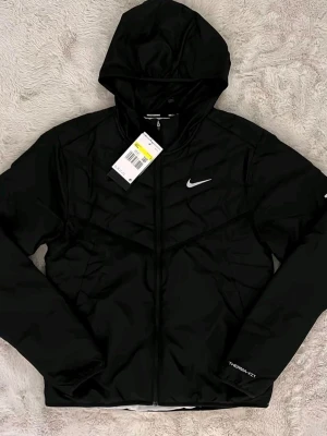 Svart Nike vindjacka med huva - Snygg svart vindjacka från Nike med huva och dragkedja framtill. Jackan har ett vitt Nike-märke på bröstet och diskret Thermasphere-tryck på ärmen. Lätt vadderad design och sportig look, perfekt för blåsiga dagar.